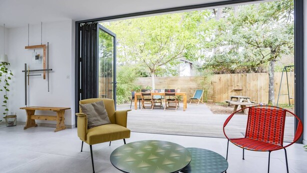 Ein modernes Interieur mit Glastür, die zu einer Terrasse führt, einer Holzbank, einem gelben Sessel und einem roten Stuhl.
