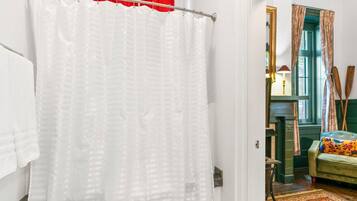 Un baño con cortina blanca de ducha, toallero y puerta que da a una habitación con un sillón verde, una lámpara y una ventana con cortinas.