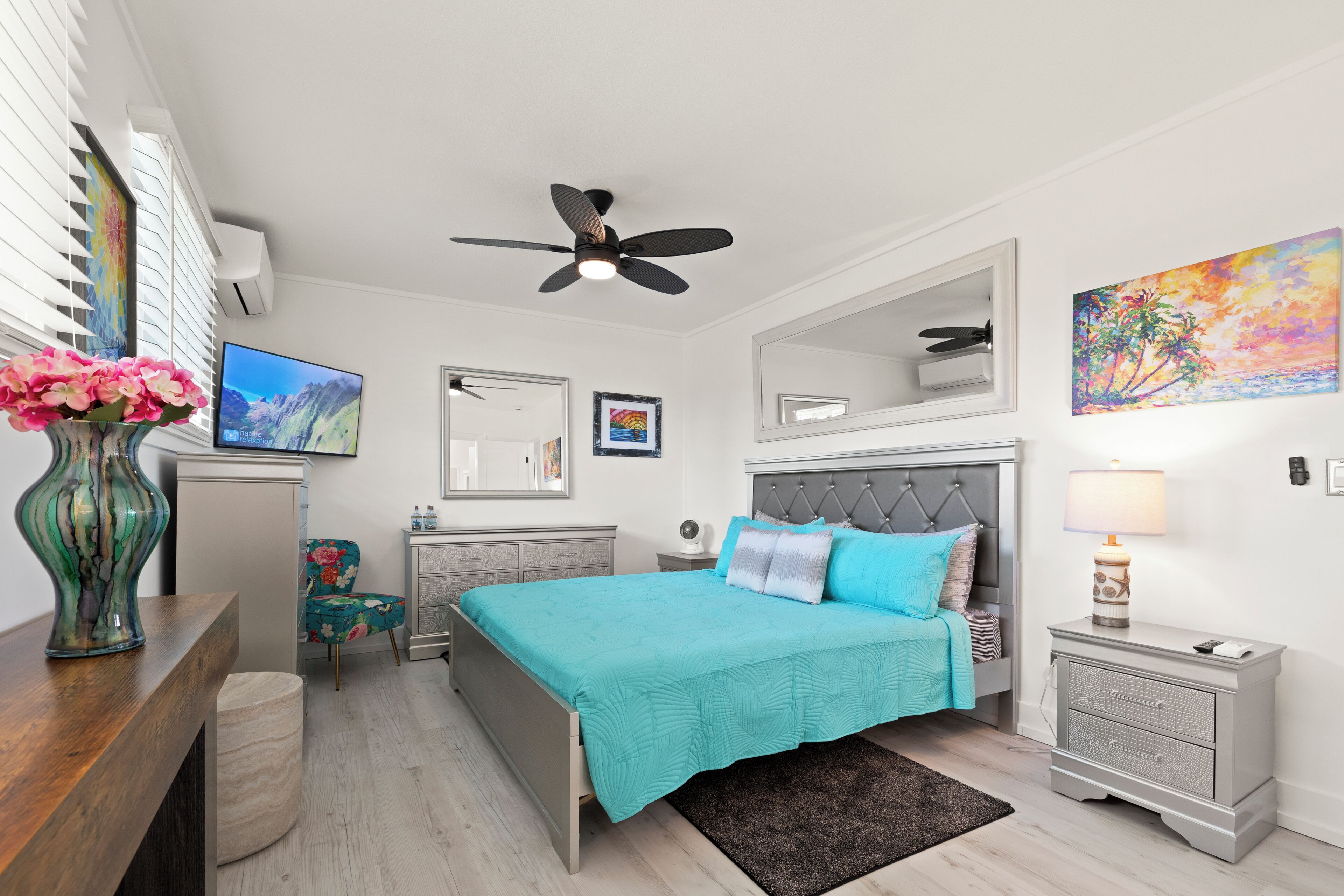 A bedroom with a bed, a nightstand, a dresser, a mirror, a ceiling fan, a TV, and a vase with flowers.