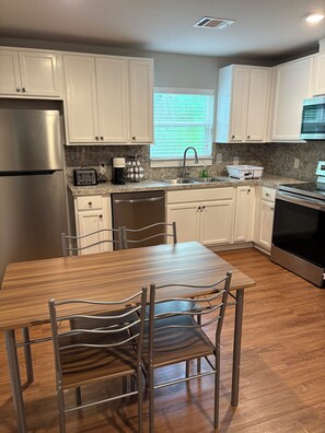 Una cocina moderna con armarios blancos, electrodomésticos de acero inoxidable, encimera de granito y un comedor con mesa de madera y sillas metálicas.
