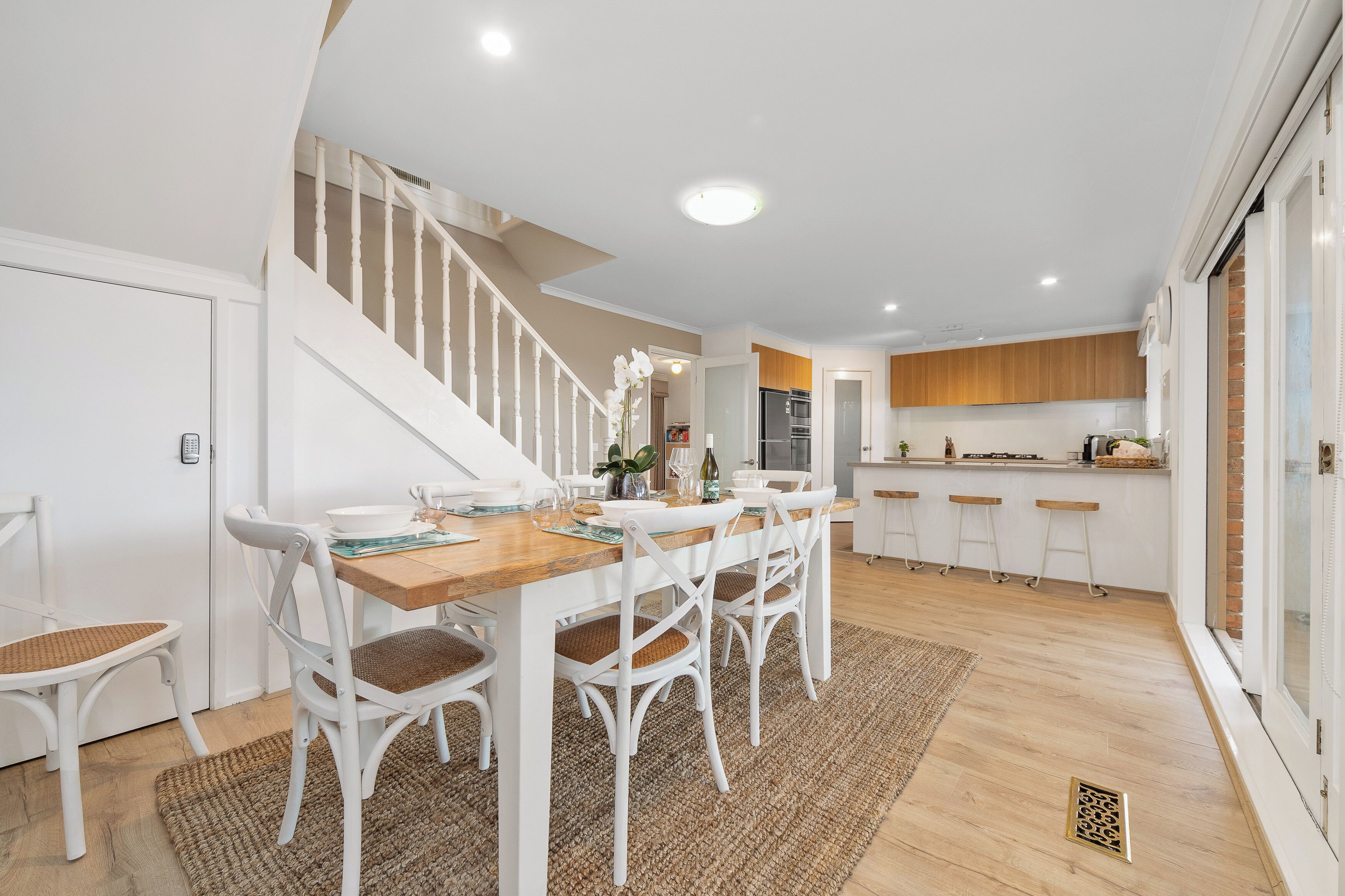 A dining area with a wooden table and white chairs, a staircase, and a kitchen with wooden cabinets.