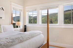 Un dormitori amb un llit amb cortines, grans finestres que donen a una vista de muntanya, una taula de nit amb una làmpada i una obra d'art penjada a la paret.