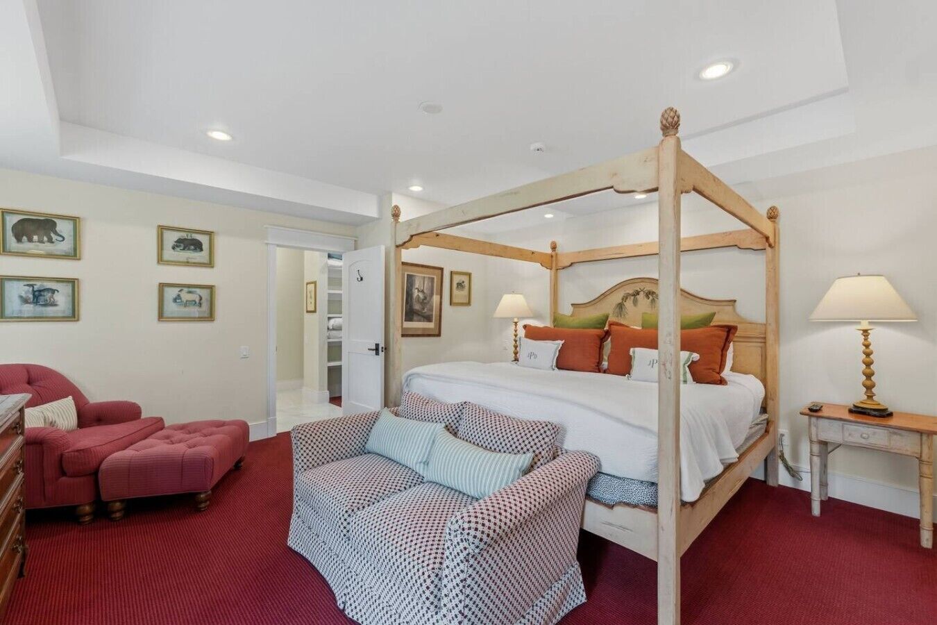 Ein Bett mit Baldachin, dessen Gestell aus Holz besteht, ein roter Teppichboden, ein roter Ohrensessel, ein gemustertes Sofa und ein Nachttisch mit Lampe.