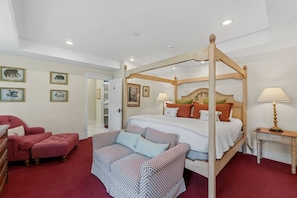 Una cama con dosel de estructura de madera, un suelo cubierto de alfombra roja, un sillón rojo, un sofá estampado y una mesita de noche con una lámpara.