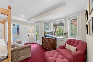 Una habitación con cama, un sofá rojo, cómoda de madera, televisor y ventanas amplias con vistas a zonas verdes.
