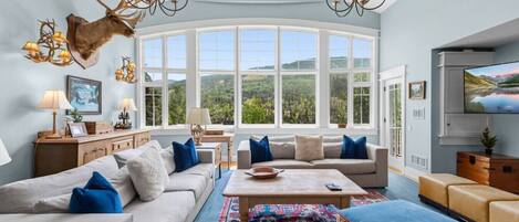 A spacious living room with a large window, a chandelier, and a mounted television.