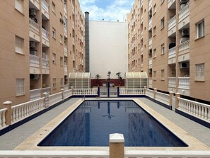A swimming pool surrounded by beige apartment buildings with balconies, a clear sky, and a central building with a white facade. - Comfort and sunshine: apartment near beach, center and community pool (Torrevieja)