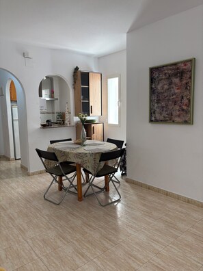 A dining area with a table set for four, a kitchen in the background, and a large abstract painting on the wall. - Comfort and sunshine: apartment near beach, center and community pool (Torrevieja)