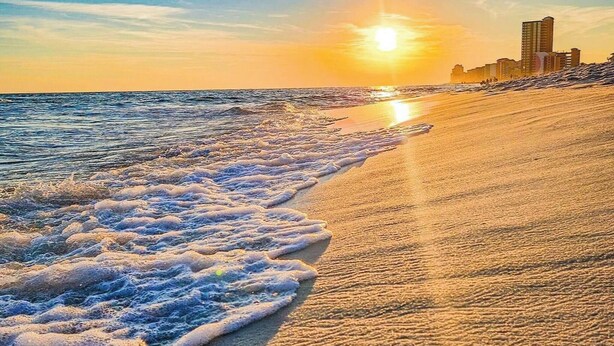 Een strand met golven, een zonsondergang en gebouwen op de achtergrond.