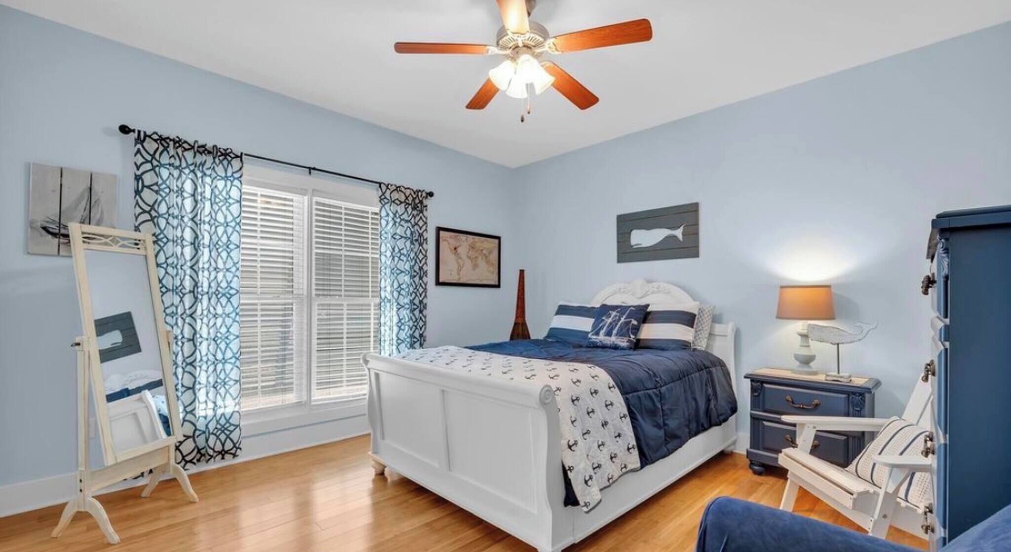 A bedroom with a bed, a ceiling fan, a bedside table with a lamp, a dresser, and a window with curtains.