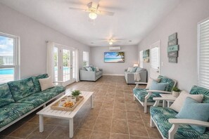 A spacious living room with a large window overlooking a beach, a ceiling fan, and a cozy seating area with a coffee table. - 7018 Rookery 3 New Listing 2 Pools 1 min to Beach (Gulf Shores)