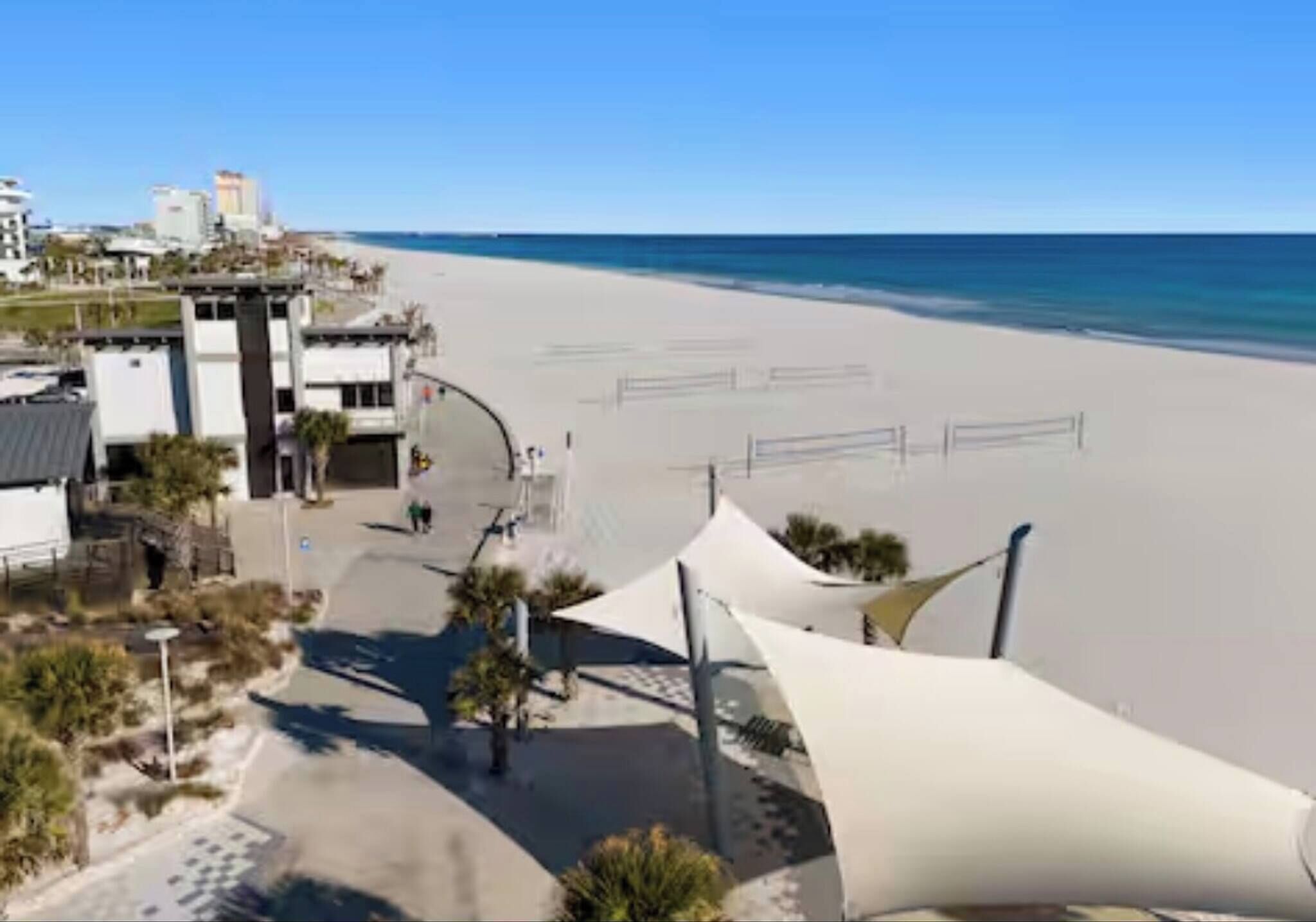 Blick auf einen Strand mit einem Weg, Strandsonnenschirmen und einem Gebäude im Hintergrund.