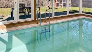 Piscina trasparente con scalette metalliche, area patio con sedie e vista su una casa attraverso ampie finestre.