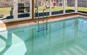 A clear swimming pool with metal ladders, a patio area with chairs, and a view of a house through large windows.