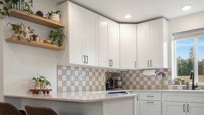 A modern kitchen with white cabinetry, a central island with bar stools, a checkered backsplash, and a window providing natural light. - Happy's Hideout, Downtown w/ Private Lawn (Ellensburg)