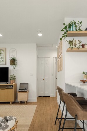 Un salon comprenant un téléviseur, une petite table, un canapé et un coin repas avec des chaises. On peut voir des plantes sur des étagères et une porte au fond.