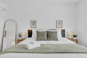 A neatly made bed with white linens, two pillows, a green throw blanket, and a folded towel. There are two bedside tables with lamps and a small potted plant.