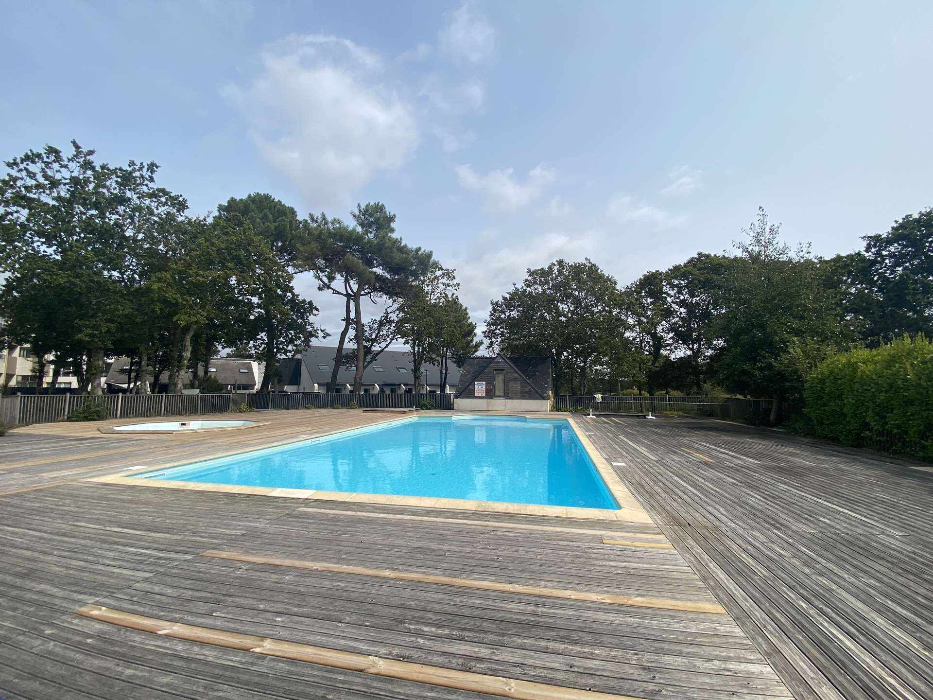 A rectangular swimming pool with a wooden deck surrounding it, set in a residential area with trees and houses in the background.