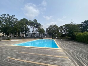 Ein rechteckiger Swimmingpool mit umlaufender Holzterrasse, eingebettet in eine Wohngegend mit Bäumen und Häusern im Hintergrund.