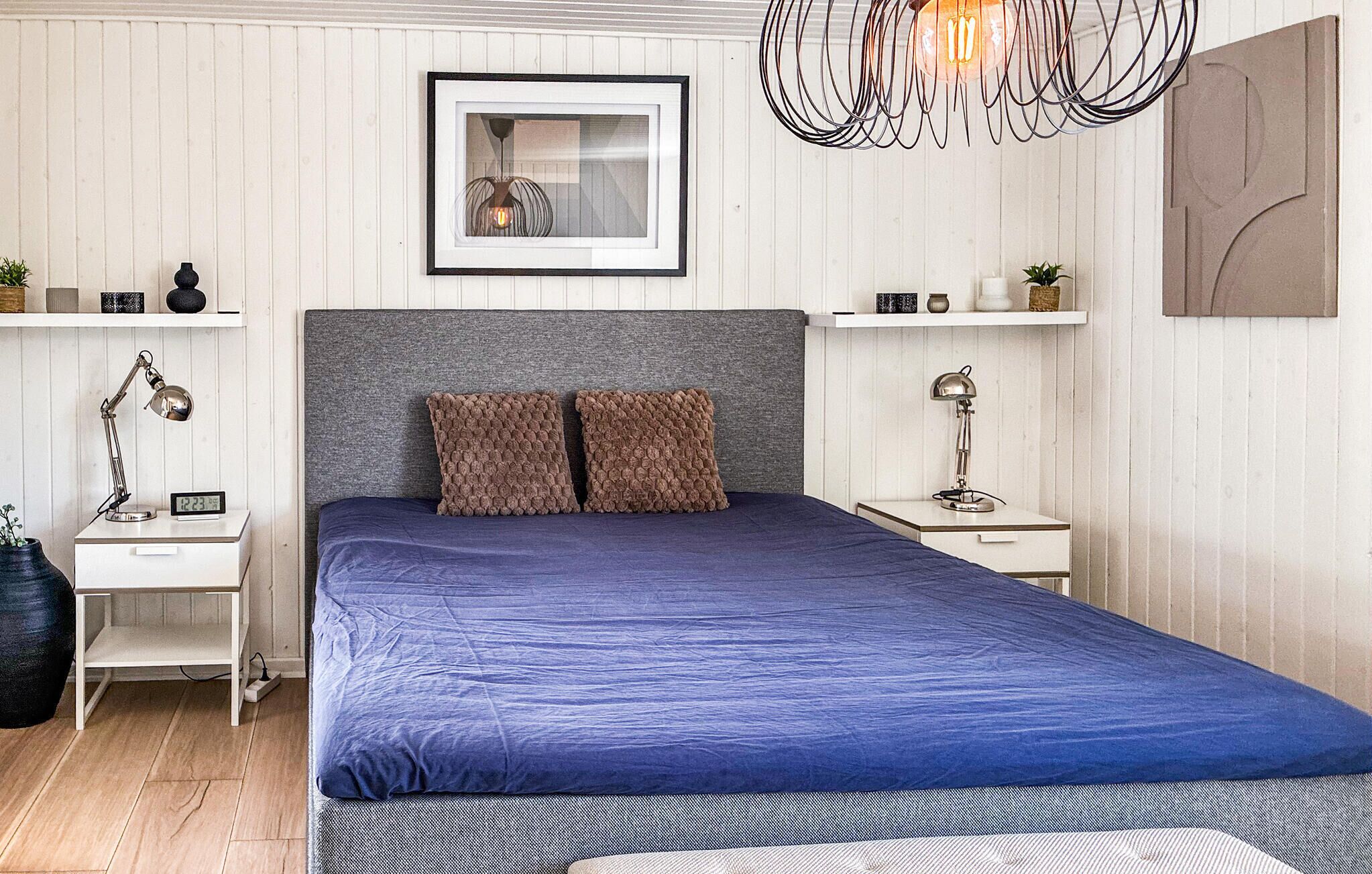 Un dormitorio con una cama, dos mesitas de noche, un cuadro colgado en la pared, una lámpara colgante y una pequeña estantería con objetos decorativos.