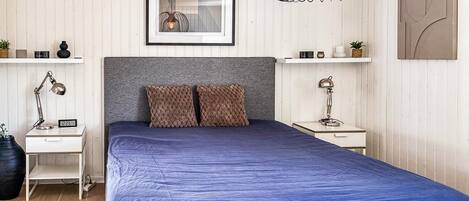 Un dormitorio con una cama, dos mesitas de noche, un cuadro colgado en la pared, una lámpara colgante y una pequeña estantería con objetos decorativos.