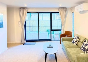 Ein Wohnzimmer mit einer Couch, einem kleinen runden Tisch und Blick durch eine Glastür auf einen Außenbereich mit Sitzgelegenheiten.