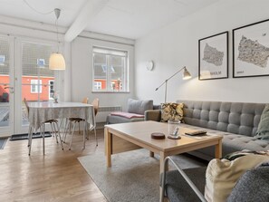 Ein modernes Wohnzimmer mit einer Couch, einem Esstisch und einem Couchtisch. Zwei Fenster mit Vorhängen, eine Lampe und gerahmte Kunstwerke sind an der Wand angebracht.