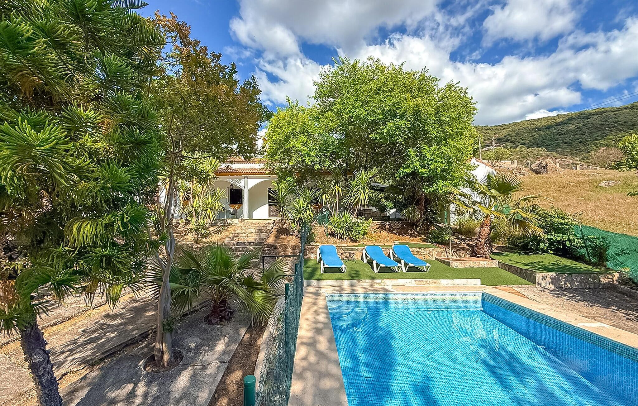 A swimming pool with blue tiles, surrounded by greenery and a white building with a tiled roof.