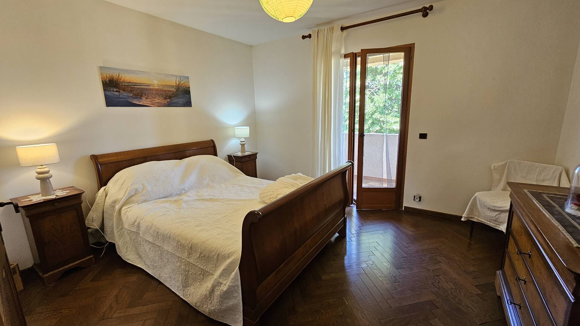 Ein Schlafzimmer mit einem hölzernen Bett, Nachttischlampen, einem Gemälde an der Wand, einer Tür zu einem Balkon und einer Holzkommode.