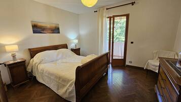 Ein Schlafzimmer mit einem hölzernen Bett, Nachttischlampen, einem Gemälde an der Wand, einer Tür zu einem Balkon und einer Holzkommode.
