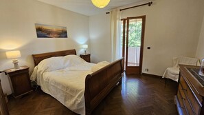 Ein Schlafzimmer mit einem hölzernen Bett, Nachttischlampen, einem Gemälde an der Wand, einer Tür zu einem Balkon und einer Holzkommode.