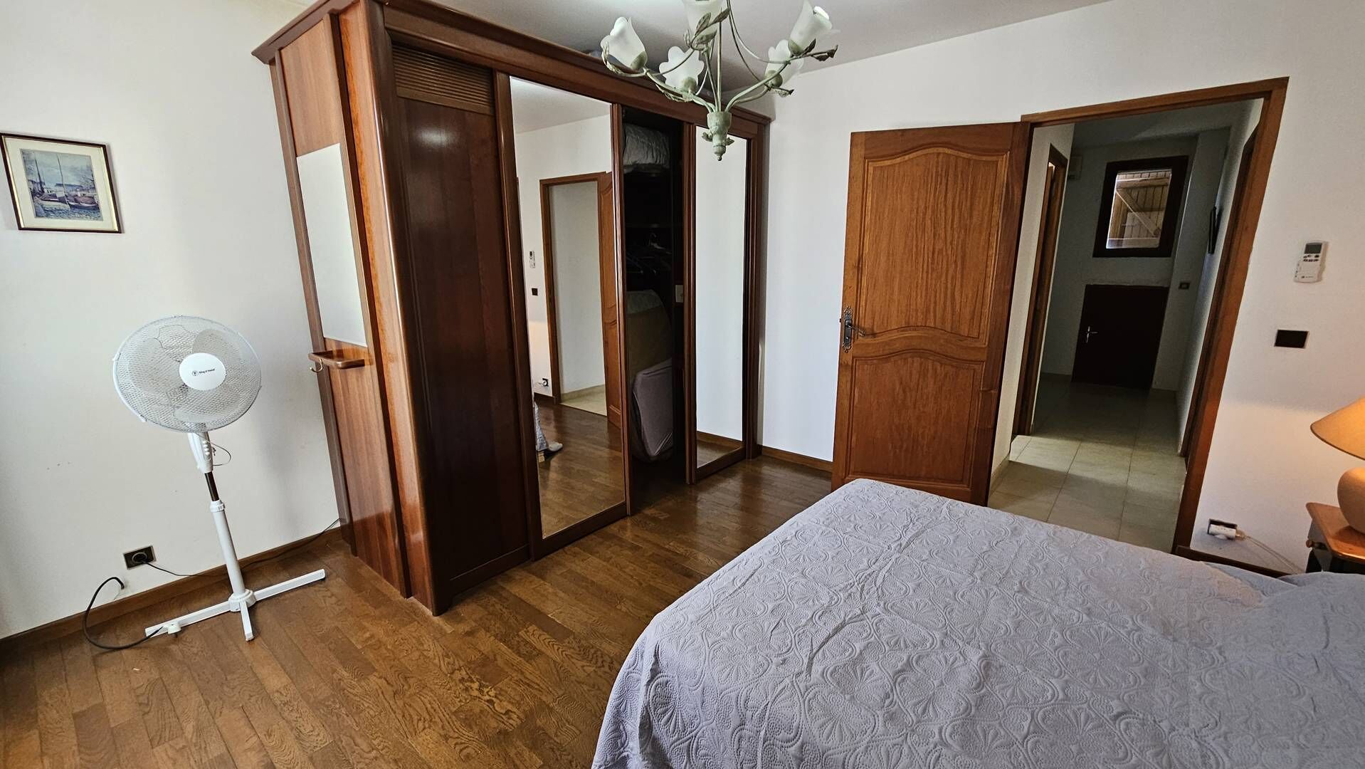 A bedroom with wooden flooring, a bed with a patterned bedspread, a standing fan, and a large mirror.