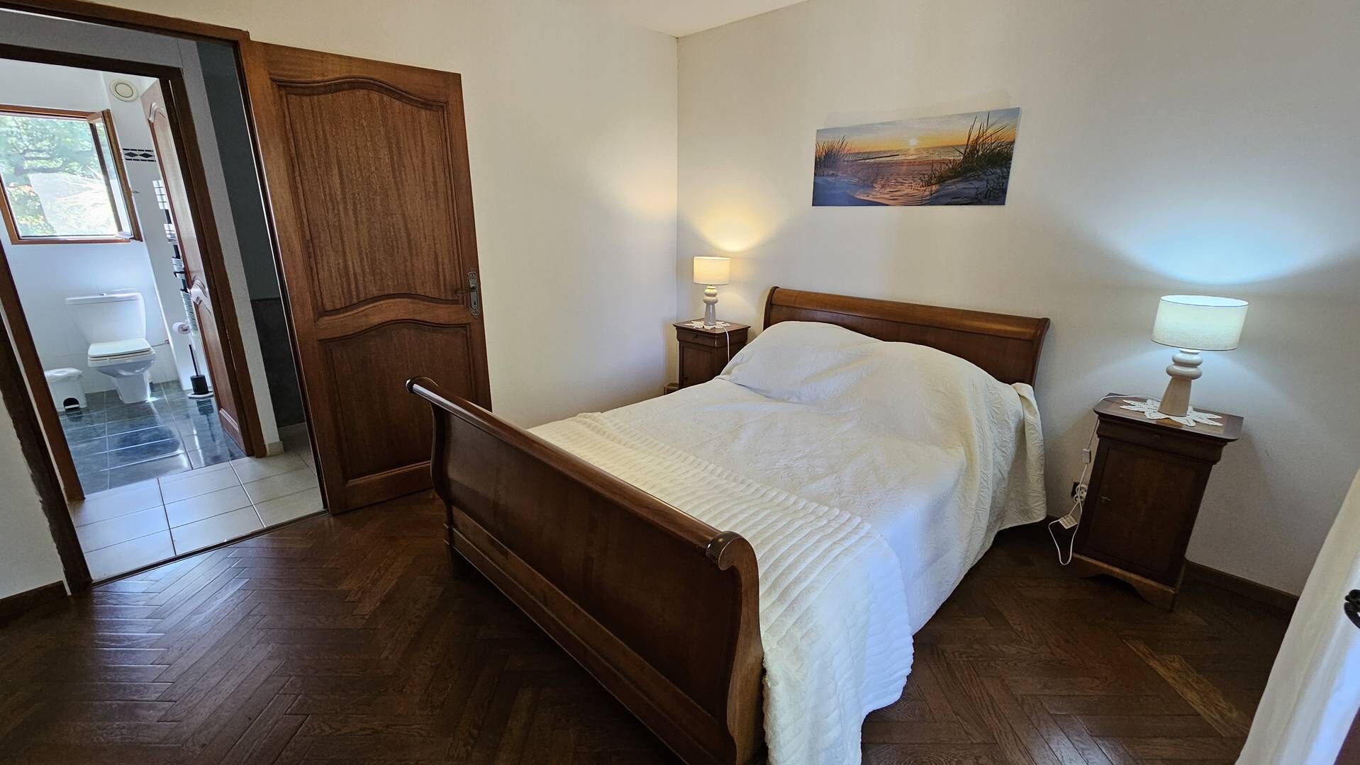A bedroom with a wooden bed, two bedside lamps, a painting on the wall, and a door leading to a bathroom.
