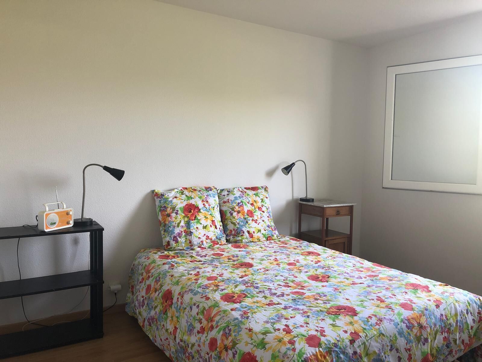 Une chambre à coucher avec une couvre-lit à motifs floraux, deux lampes de chevet, une petite table et une radio posée sur une table de nuit.