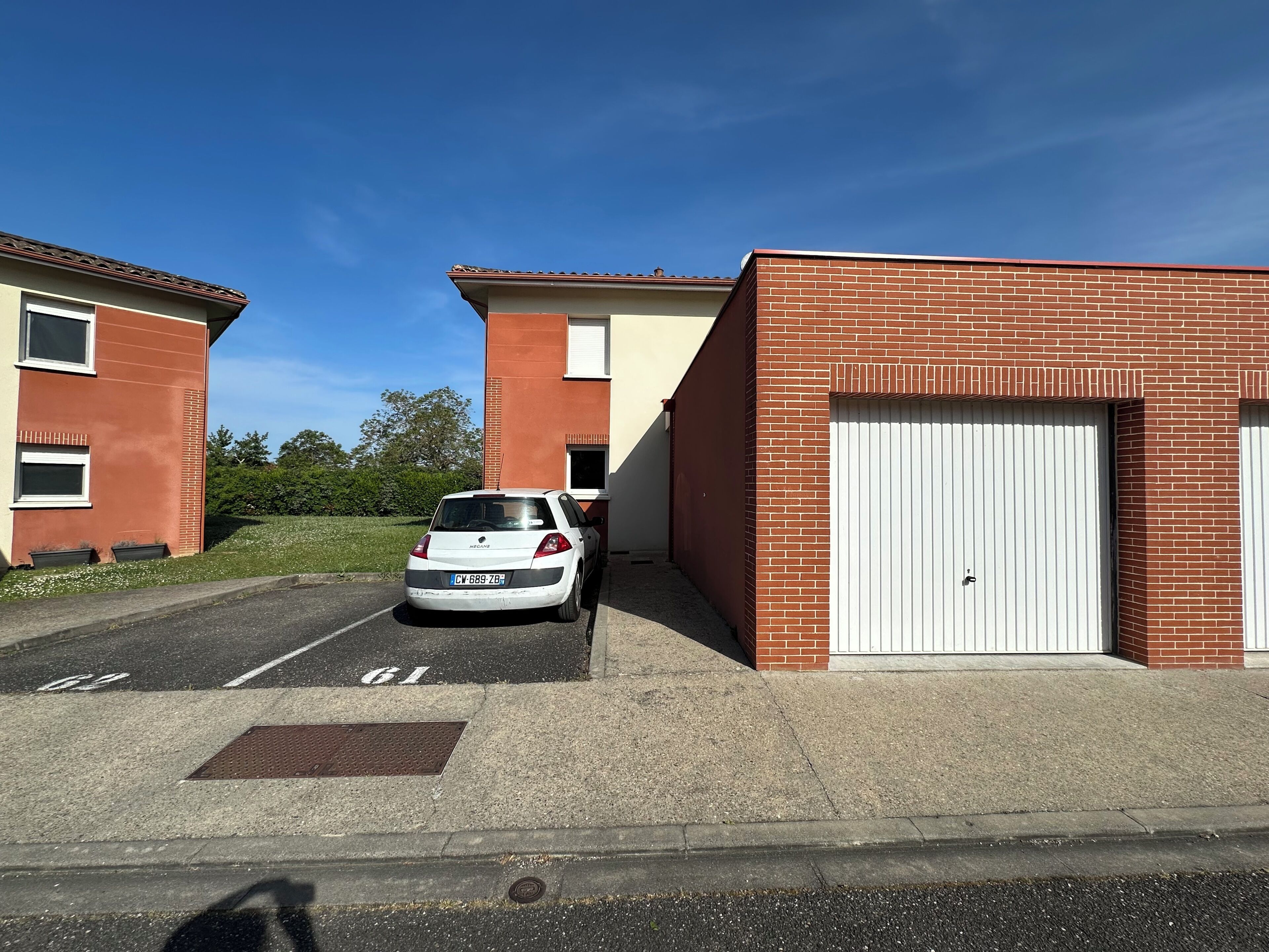 A parking area with a white car, a building with a red brick facade, and a garage door.