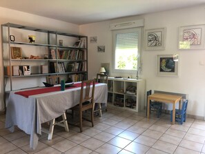 Une pièce comprenant une table à manger, des chaises, une bibliothèque, une fenêtre avec des stores et des œuvres d’art accrochées aux murs.