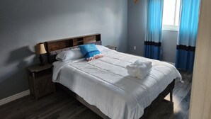 Ein ordentlich bezogenes Bett mit weißen Leinentüchern und einem blauen Kopfkissen, ein Nachttisch aus Holz mit einer Lampe und ein Fenster mit blauen Vorhängen.