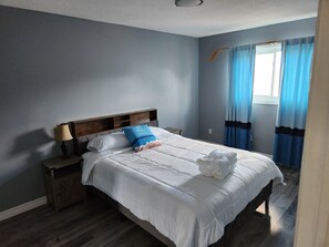 Un lit bien fait, avec du linge de lit blanc et un oreiller bleu, une table de chevet en bois avec une lampe, et une fenêtre avec des rideaux bleus.