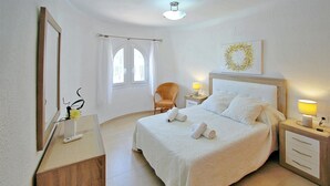 Ein ordentlich eingerichtetes Hotelzimmer mit Bett, Nachttischen, einem Sessel, einem Spiegel und einem Fenster mit Vorhängen.