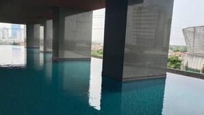 An indoor swimming pool with clear water, concrete pillars, and a view of buildings in the background. - Grand Sungkono Lagoon Caspian Apartment 1BR (Surabaya)