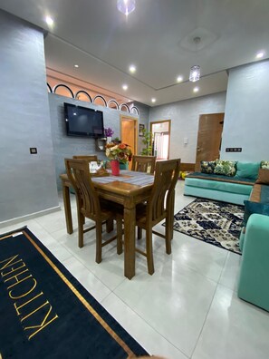 A dining area with a wooden table and chairs, a TV mounted on the wall, and a sofa in the background. - Luxury 2-bedroom apartment (Kénitra)
