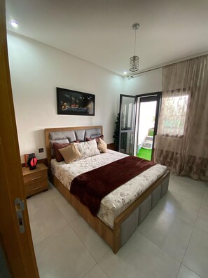 A bedroom with a large bed, a nightstand, a painting on the wall, and a view through a glass door. - Luxury 2-bedroom apartment (Kénitra)