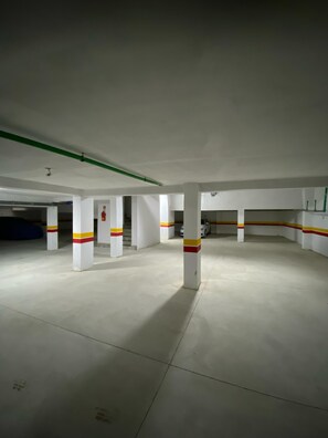 An empty parking garage with white pillars marked with red and yellow stripes, a green pipe on the ceiling, and a white car parked in the distance. - Luxury 2-bedroom apartment (Kénitra)