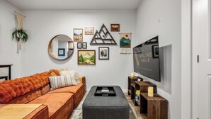 A living room with a sofa, ottoman, TV, wall art, and a plant.