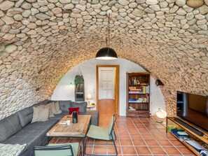 Un techo de piedra arqueado, un rincón acogedor con sofá y sillones, una mesa de centro de madera, una estantería y un televisor.