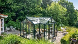 Una estructura de invernadero con techo y paredes de vidrio, rodeada por un jardín con diversas plantas y muebles de exterior.