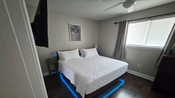 Una camera da letto con un letto, una ventola a soffitto, una finestra con tende e un'opera d'arte appesa al muro.