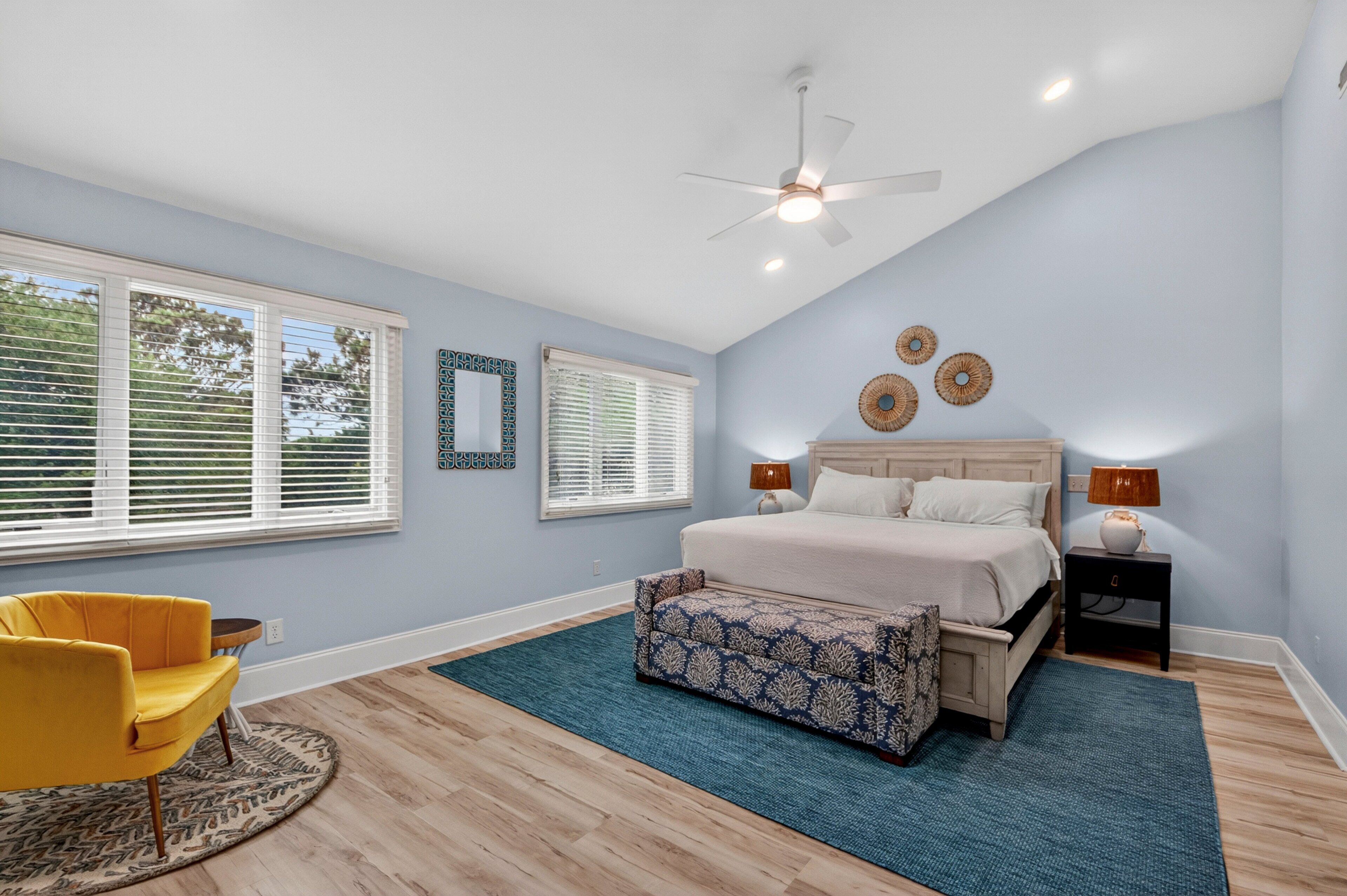A bedroom with a bed, a ceiling fan, a nightstand, a lamp, a chair, and a window with blinds.