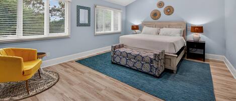 A bedroom with a bed, a ceiling fan, a nightstand, a lamp, a chair, and a window with blinds.