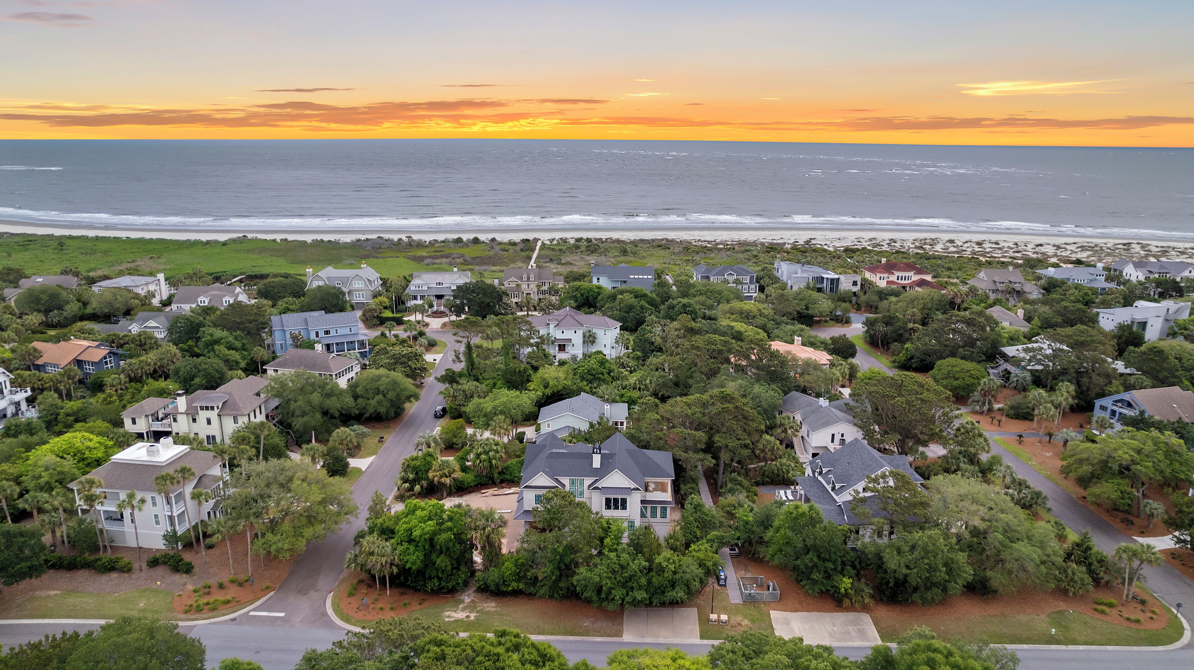 A residential area with houses surrounded by greenery and a view of the ocean at sunset.